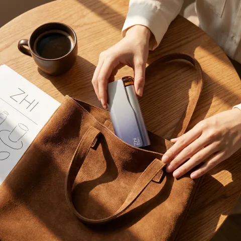 Hands slipping the Zhithera D1 waterless diffuser into a brown suede tote beside a coffee cup