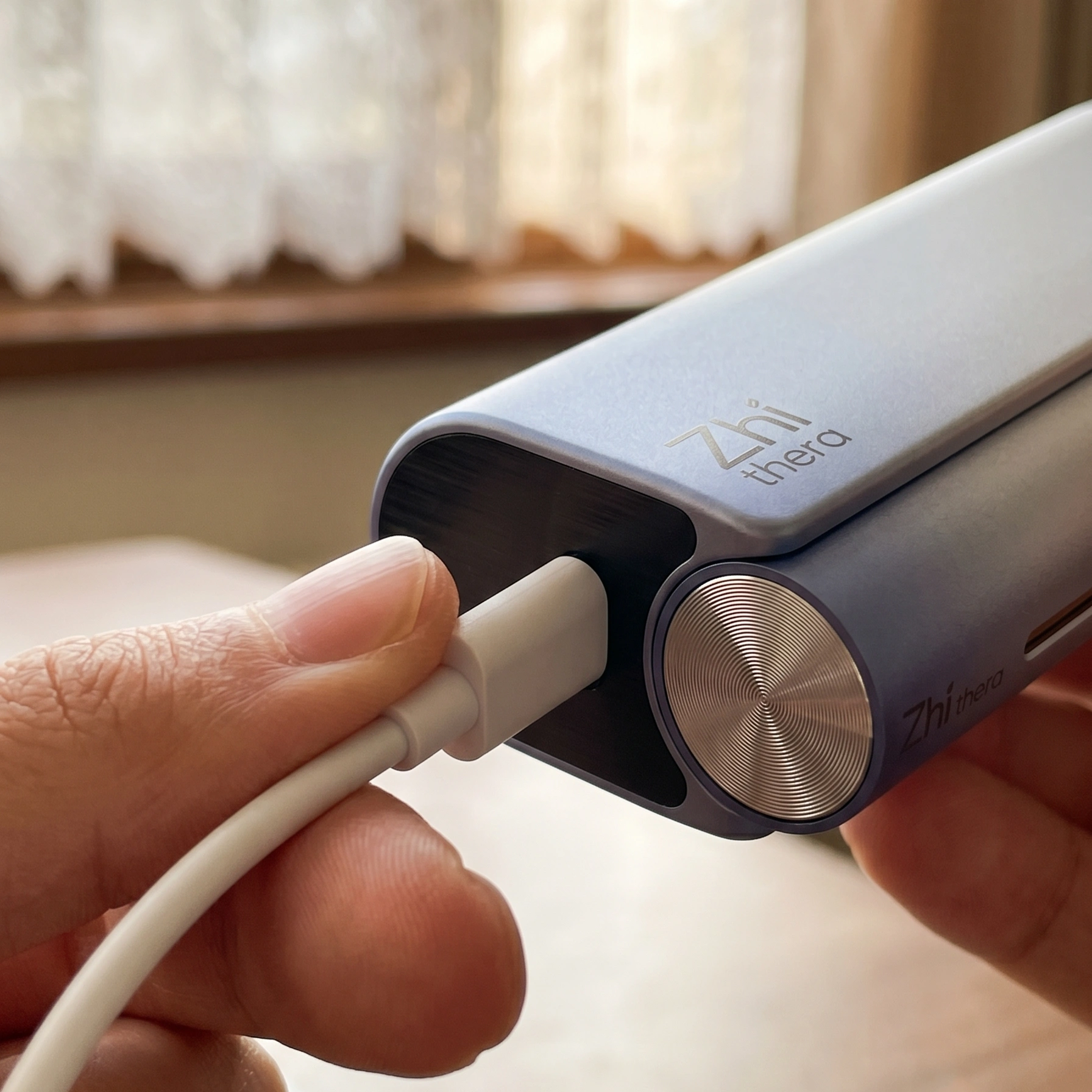 A hand plugs a white USB-C cable into a light blue-gray Zhithera portable device held near a sunlit window with sheer curt…