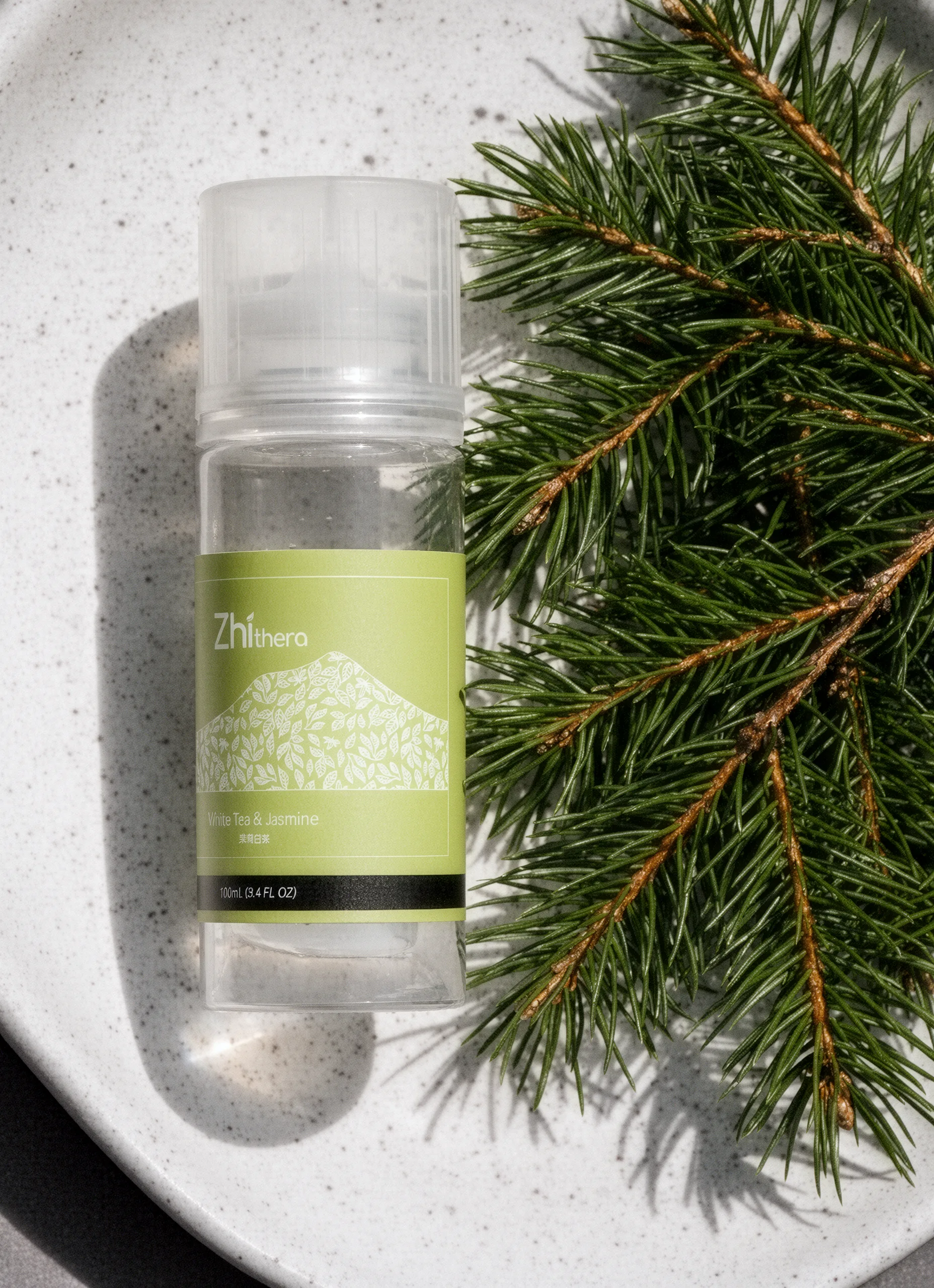 A clear bottle of Zhithera essential oil with a sage green label on a speckled white ceramic plate beside fresh pine
