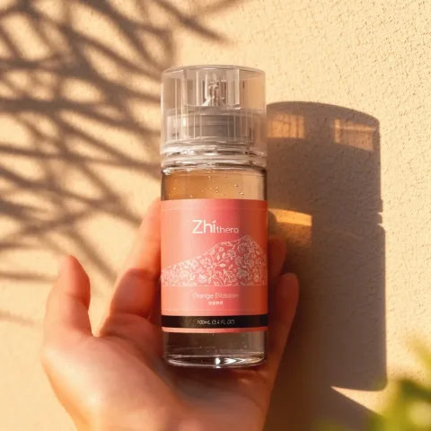 Hand holding a Zhithera Orange Blossom 100 ml fragrance oil bottle in palm-leaf sunlight